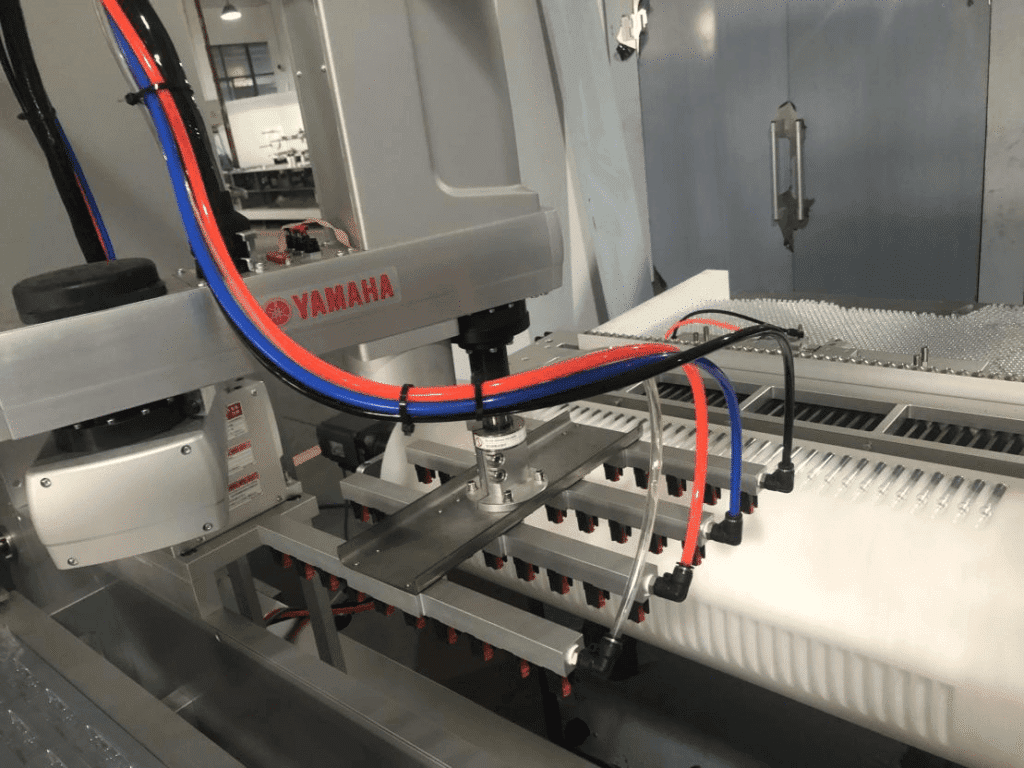 Yamaha robotic arm loading system with vacuum suction picking up ampoules or vials on a pharmaceutical cartoning line.